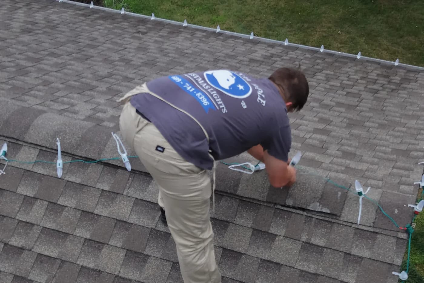 Seen from above, an installer bent over the ridge clipping lights, the North Pole mark across his back