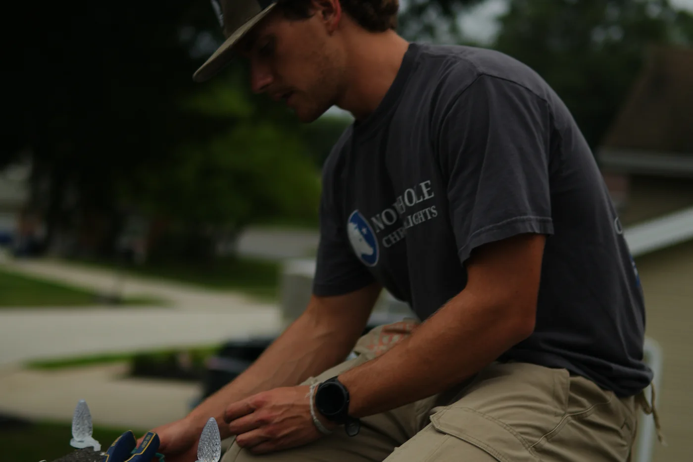 A North Pole installer sitting on the roof edge clipping C9 bulbs onto a strand, the logo on his shirt