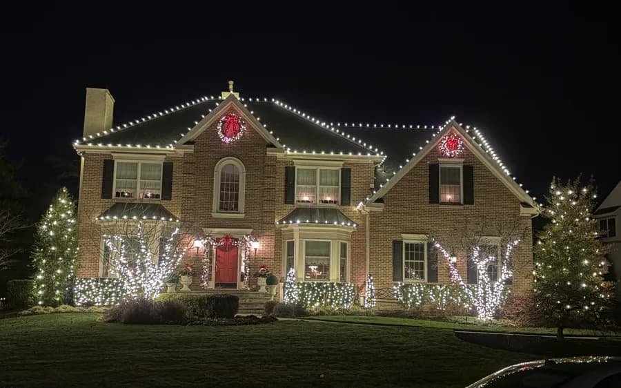 A brick home with lit wreaths in the upper windows and trees wrapped either side