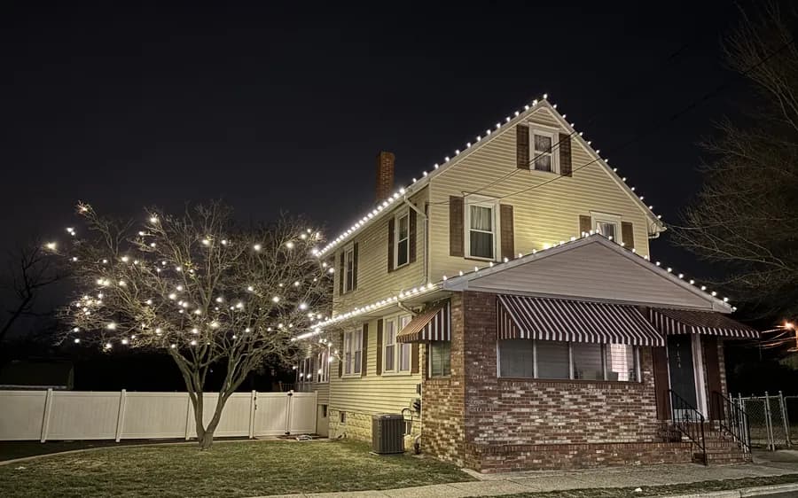 A pale clapboard home with a fully lit tree in the front yard