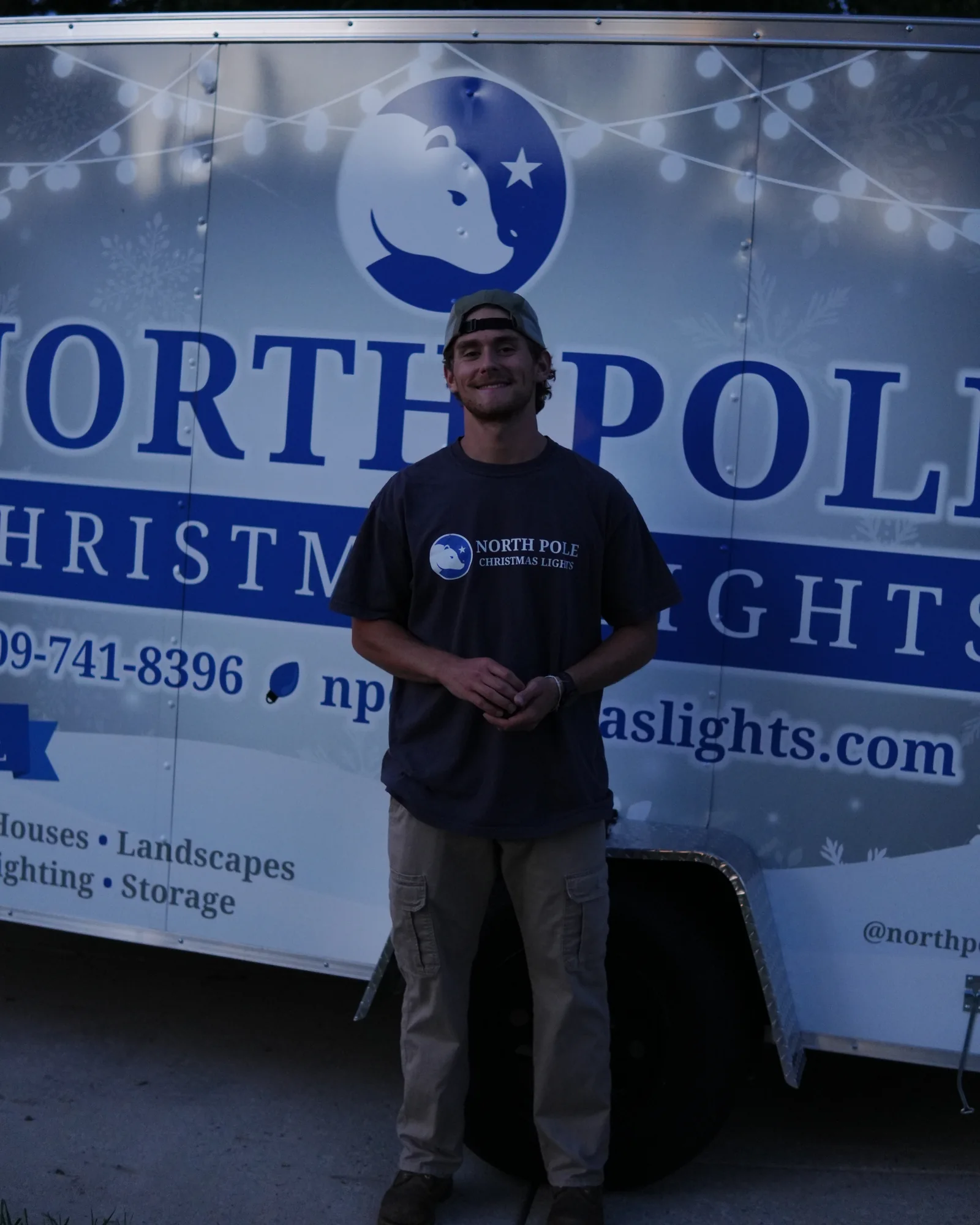 A North Pole installer smiling in front of the branded trailer at dusk, the bear mark and wordmark behind him