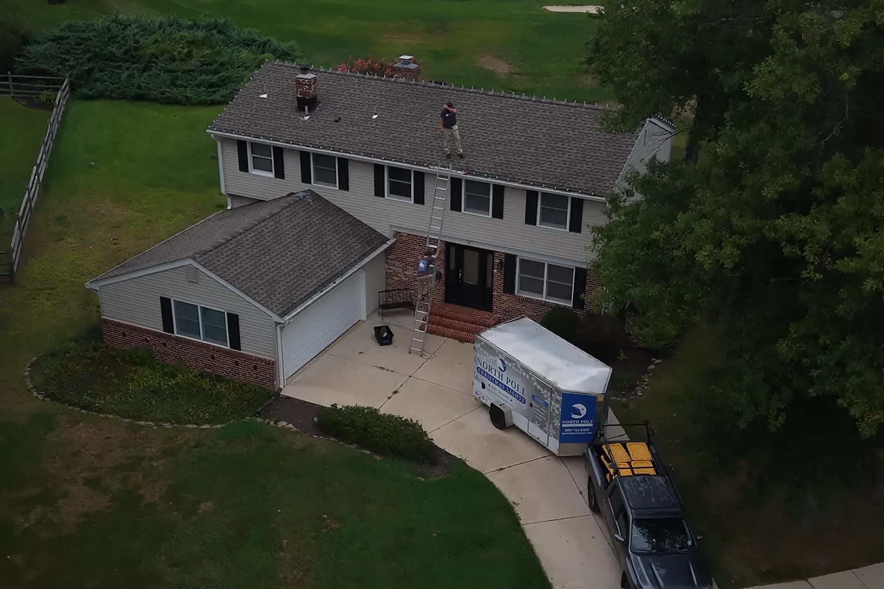 Seen from a drone, the North Pole truck and branded trailer in a driveway while installers work from the roof and a ladder