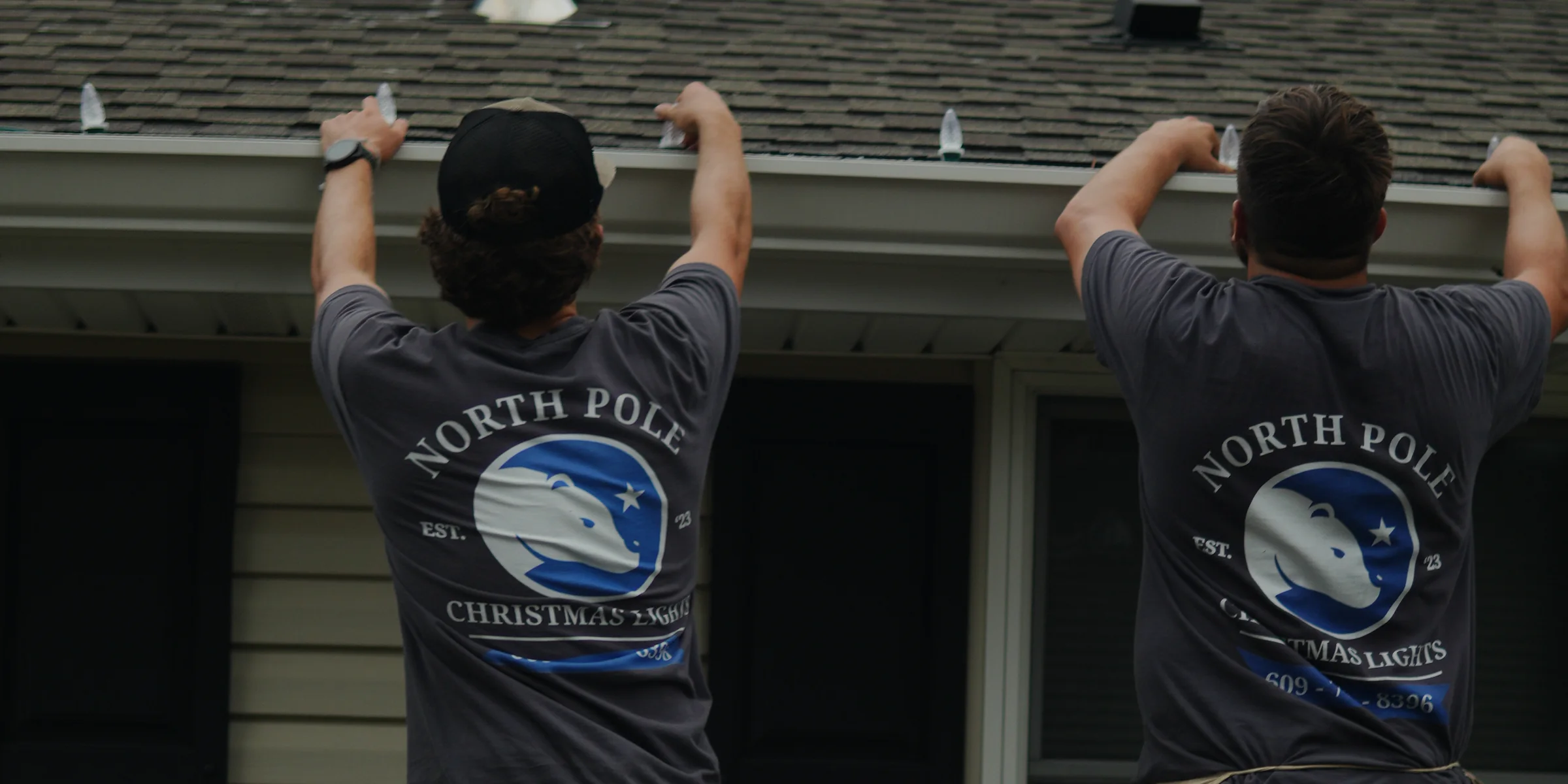 North Pole installers on a roof in branded shirts, the bear mark and phone number across the back