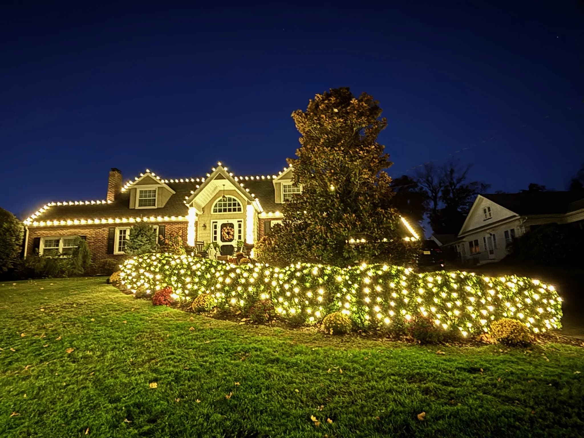 A home at blue hour with warm white lights along the roofline, the bushes out front lit and a tree beside the house