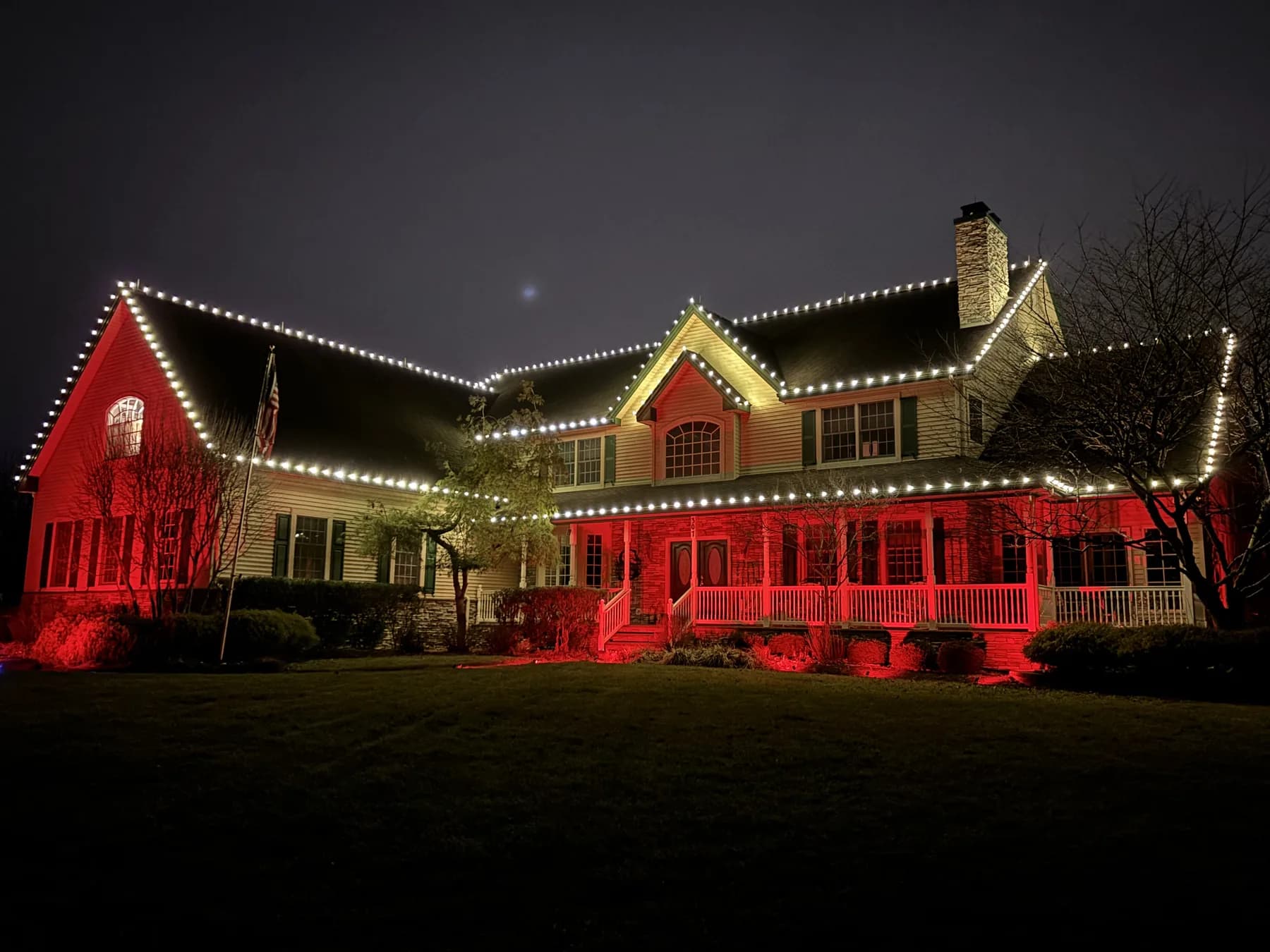 A two-story home at night with warm white rooflines and red light washed across the porch and front wall