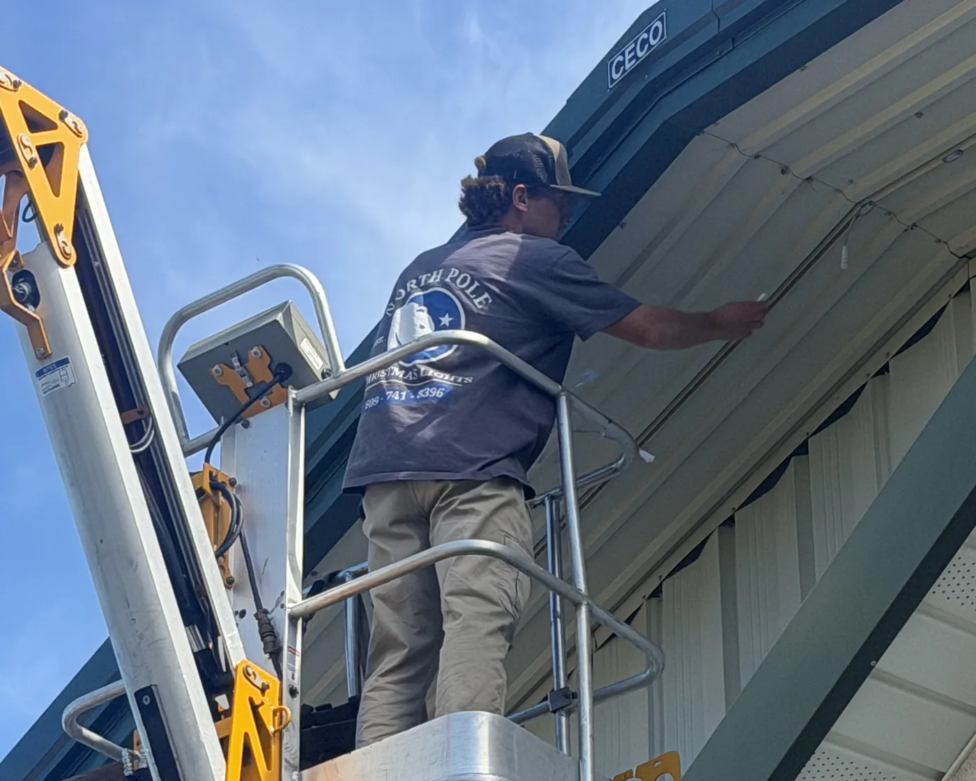 A North Pole installer on a boom lift fitting a permanent lighting track under the eave of a commercial building