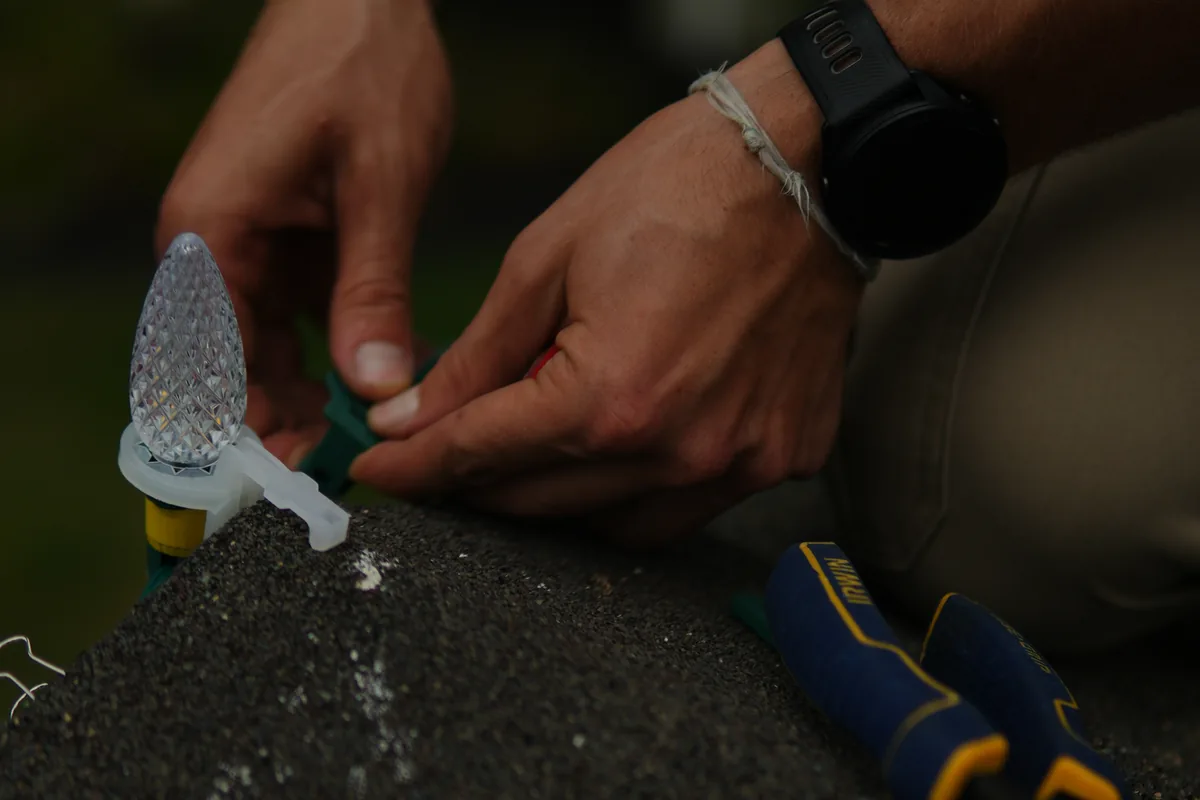 Two hands fitting a C9 bulb into its clip on the ridge, pliers resting on the shingle beside them